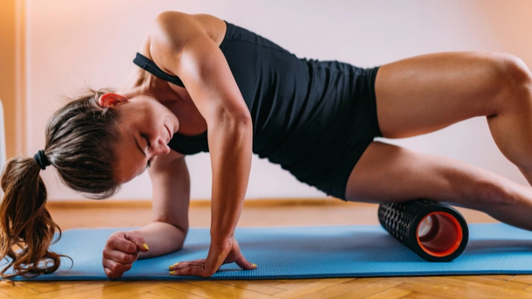 A woman with a ponytail is shown on a blue yoga mat, she is wearing a black top and black shorts and is using a foam roller to massage her leg muscles, the foam roller is positioned underneath her leg, and she is using her arms to support her upper body, the woman appears focused on the task at hand, with her eyes closed and a slight expression of concentration on her face.