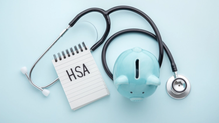 A blue ceramic piggy bank, a stethoscope, and a notepad with letters 'HSA' on it is displayed on top of a light blue matte surface.